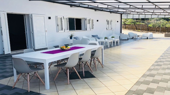 Un patio al aire libre con una mesa y sillas blancas, una zona cubierta con pérgola y vistas a un jardín.