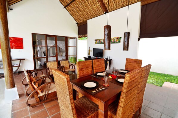 A dining area with a wooden table and wicker chairs, a television, and a large window with a view of greenery.