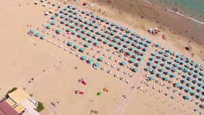 Une vue aérienne d’une plage, avec des rangées de parasols et de chaises longues, ce qui indique qu’il s’agit d’un complexe hôtelier balnéaire.