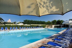 Una piscina con sedie e ombrelloni blu, circondata da vegetazione e edifici.