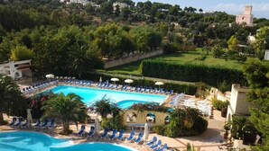 Un complexe hôtelier doté d’une grande piscine entourée de chaises longues et de parasols, le tout situé dans un cadre verdoyant avec une végétation luxuriante et des collines en arrière-plan.