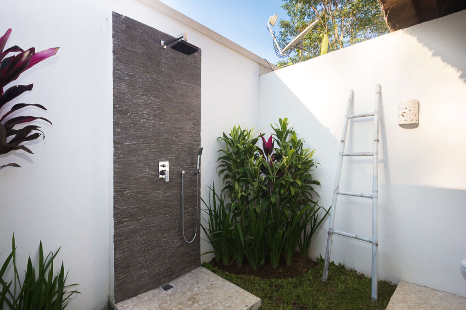 Un área de ducha exterior moderna con pared de piedra, escalera metálica y abundante vegetación.