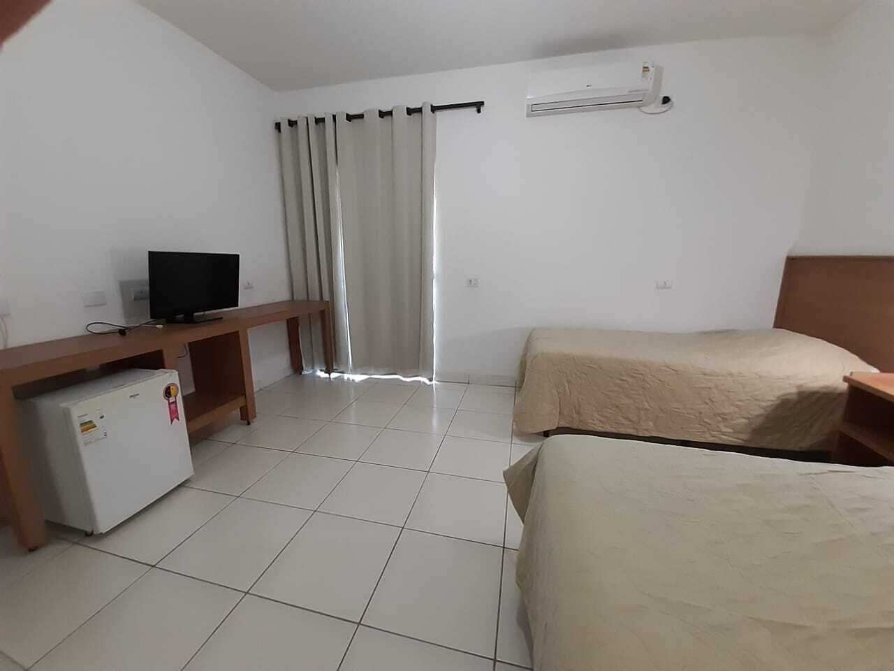 A hotel room with two beds, a TV, a small fridge, and a window with curtains.