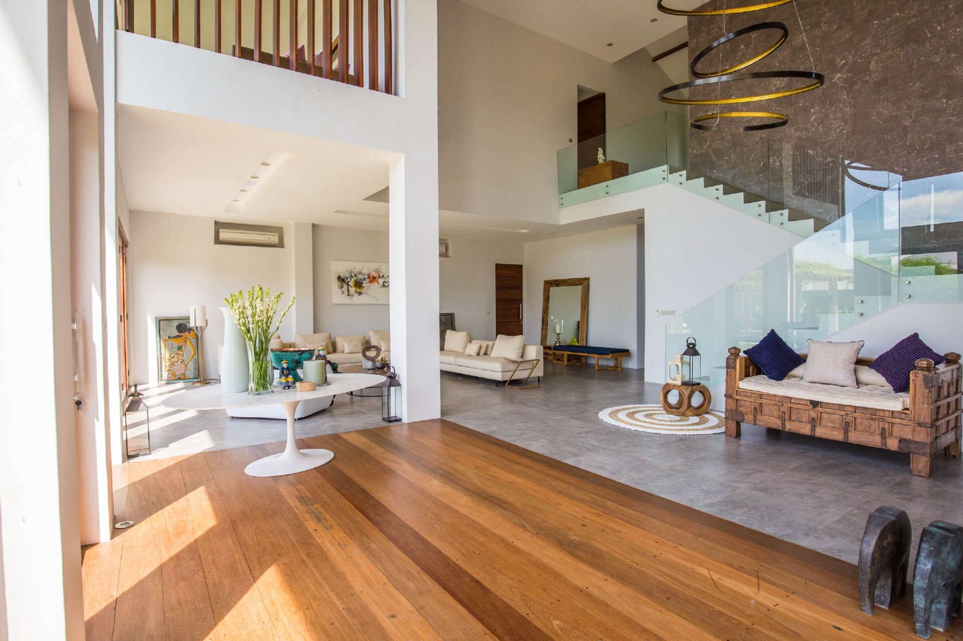 Amplia sala de estar con piso de madera, un sofá blanco, un banco de madera y una escalera con barandillas de vidrio.