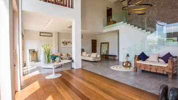 Amplia sala de estar con piso de madera, un sofá blanco, un banco de madera y una escalera con barandillas de vidrio.