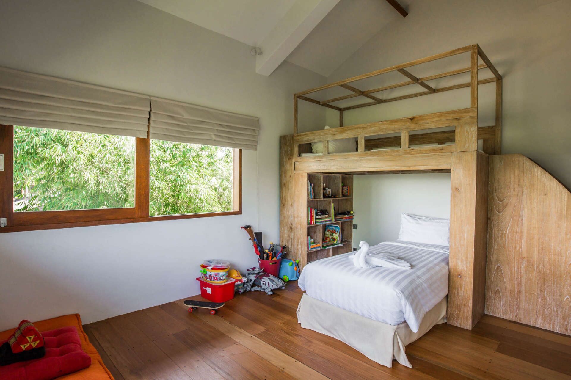 Una cama individual con estructura de madera, un escritorio pequeño y una ventana con vista a follaje.
