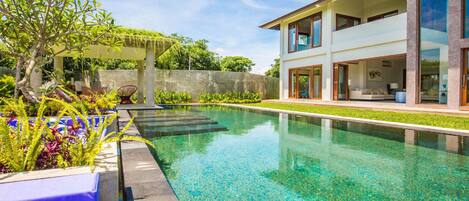 A modern house with a swimming pool, a covered outdoor seating area, and lush greenery.