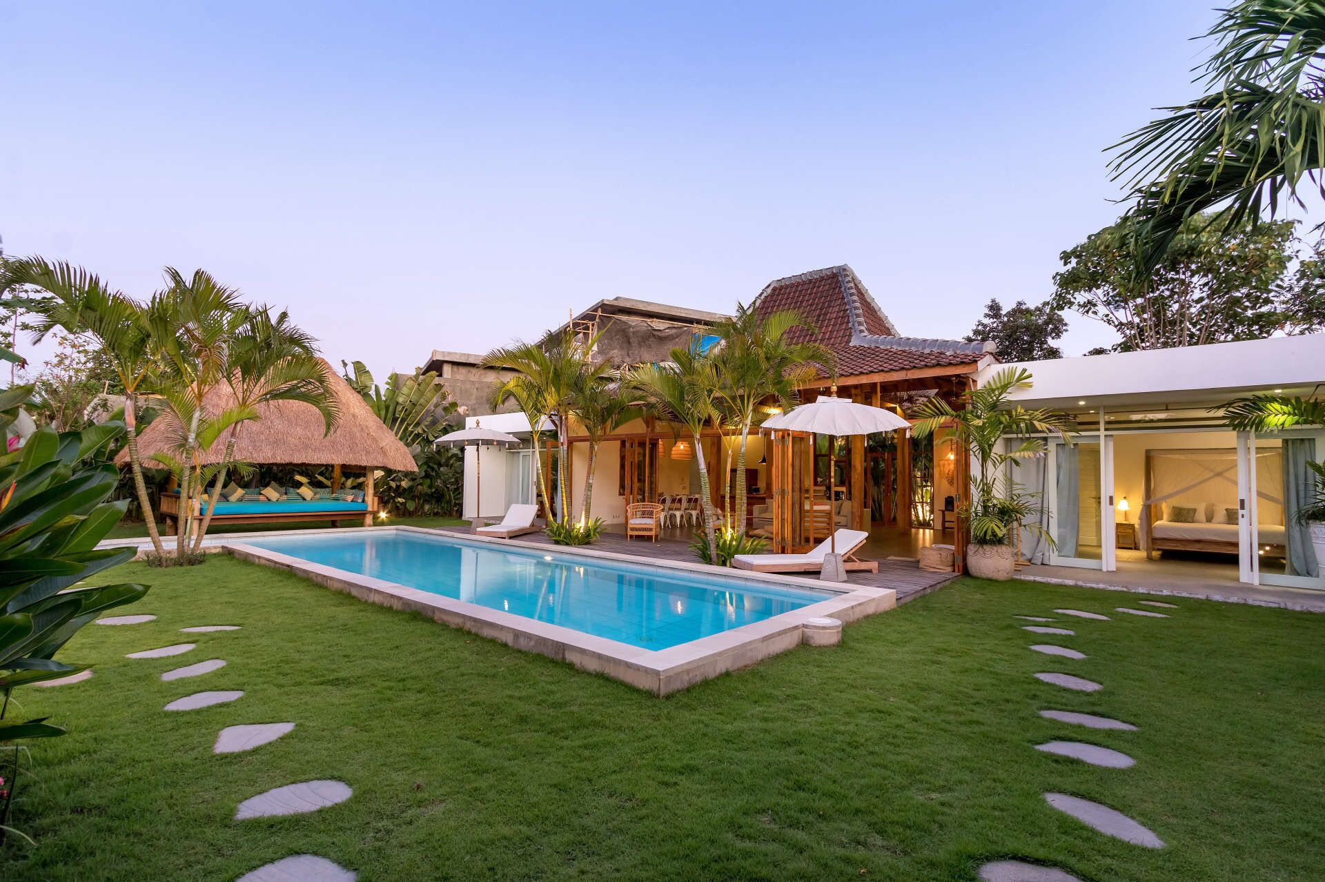 Una piscina rodeada por un césped cuidado, piedras para pisar, una construcción con techo de paja y una casa moderna con asientos exteriores y sombrillas.