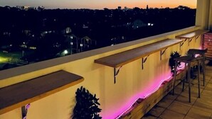 A balcony with a view of a cityscape at dusk. There are wooden benches and a small table.