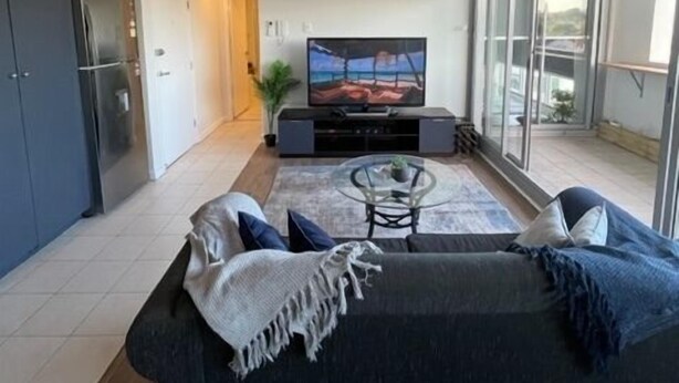 A modern living room with a sofa, a coffee table, and a TV. There is a kitchen area with a refrigerator and cabinets. The room has large windows and a balcony.