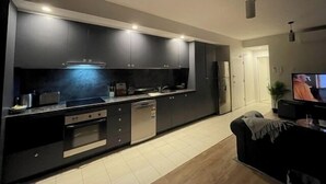 A modern kitchen with dark cabinetry, a built-in oven, and a dishwasher. There is a dining area with a black sofa and a TV mounted on the wall.