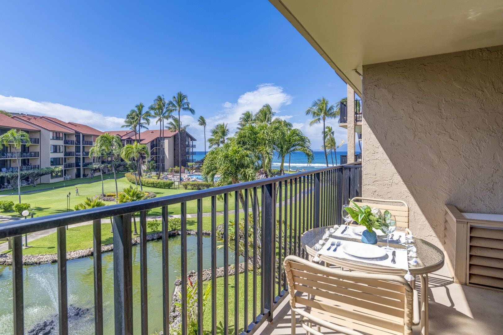 A balcony with a table set for dining, overlooking a pool and a golf course with palm trees and a building in the background.