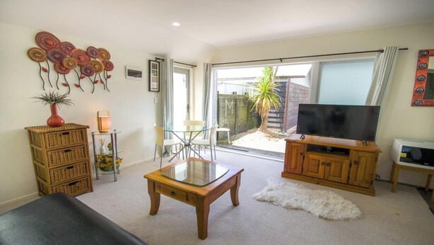 A living room with a glass table, wicker furniture, a flat-screen TV, and a sliding glass door leading to a garden. - Beautiful & Bright Studio APT (Auckland)