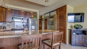 A modern kitchen with wooden cabinets, stainless steel appliances, a granite countertop, and a bar area with two stools. - Newly upgraded kitchen with oceanfront views (Lahaina)