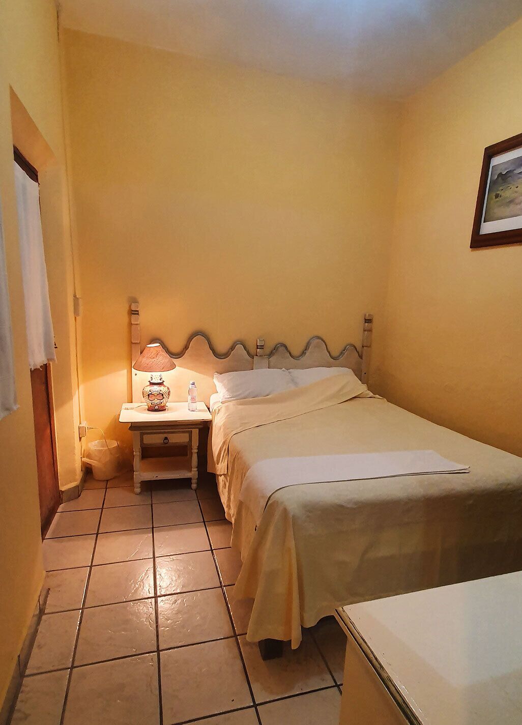 A small, neatly arranged bedroom with a single bed, a bedside table with a lamp, and a wall-mounted picture.