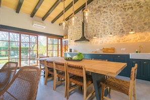 A spacious kitchen with a large wooden table and wicker chairs, stone wall backsplash, and hanging pendant lights.