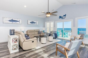 A spacious living room with a ceiling fan, a large window overlooking the ocean, and a view of the beach.