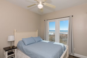 Quarto com cama, mesa de cabeceira, abajur, ventilador de teto e janela com vista para o oceano.