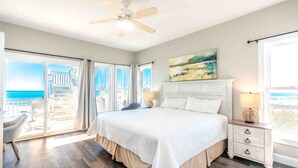 A bedroom with a large bed, a ceiling fan, a nightstand, and a view of the ocean through the window.