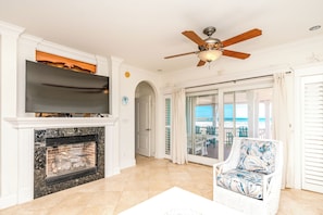 A living room with a fireplace, a television, a ceiling fan, and a view of the ocean through a glass door.