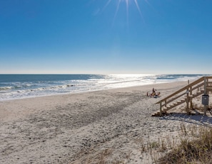 A beach with a wooden staircase leading down to the sand, a clear sky, and the ocean in the background.