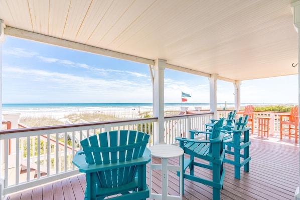 A covered outdoor area with a view of the beach, featuring Adirondack chairs and a small table.