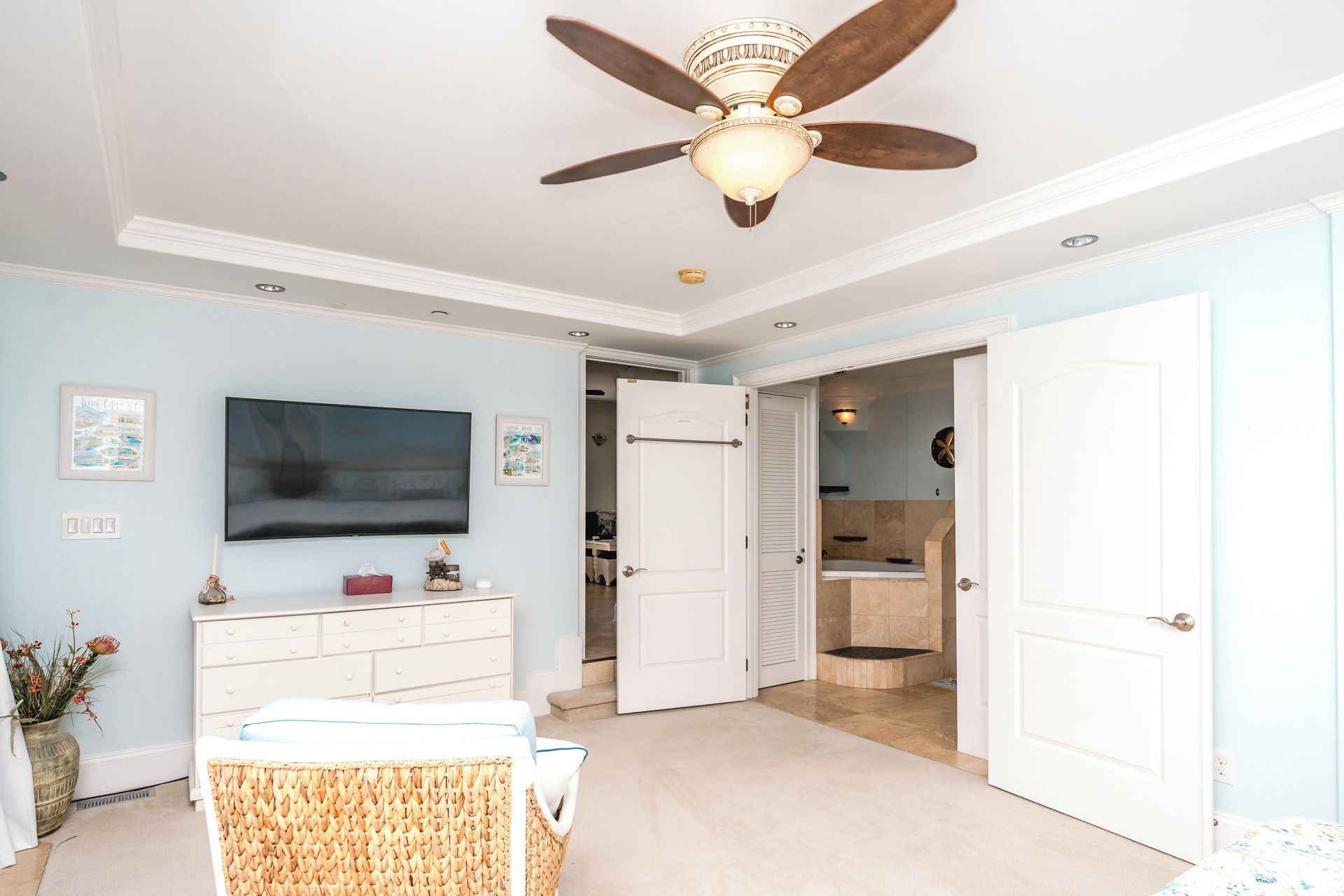 A room with a ceiling fan, a flat-screen TV, a white dresser, a wicker chair, and a door leading to a bathroom.