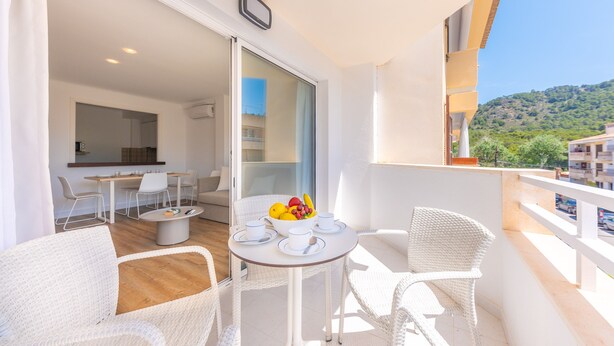 Uma varanda com móveis de vime branco, uma mesa com uma fruteira e vista para uma encosta.