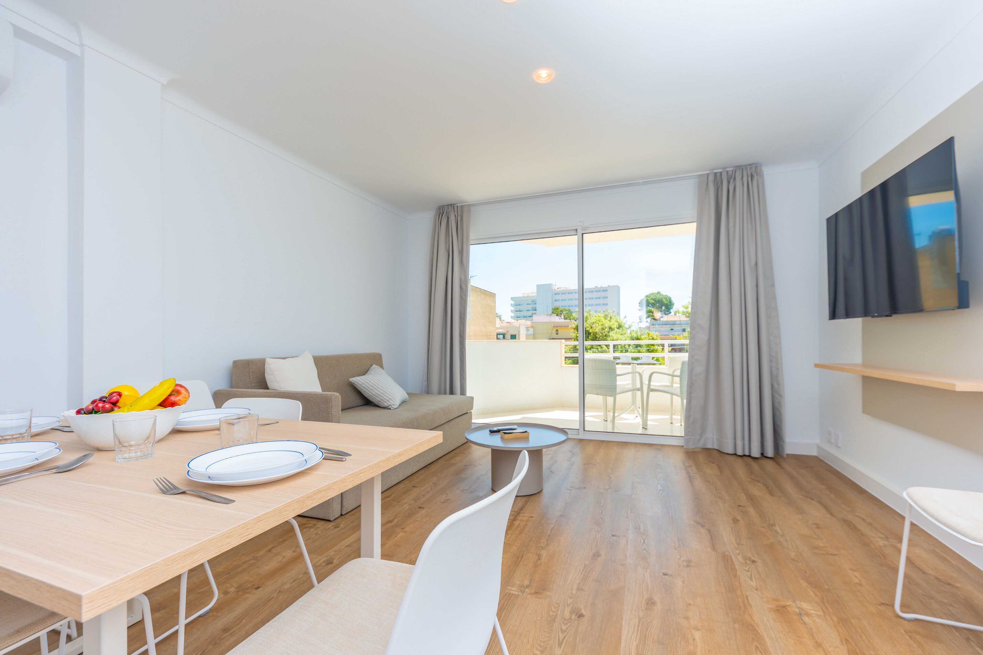 A modern living room with a dining area, a sofa, a TV mounted on the wall, and a balcony with a view of the city.