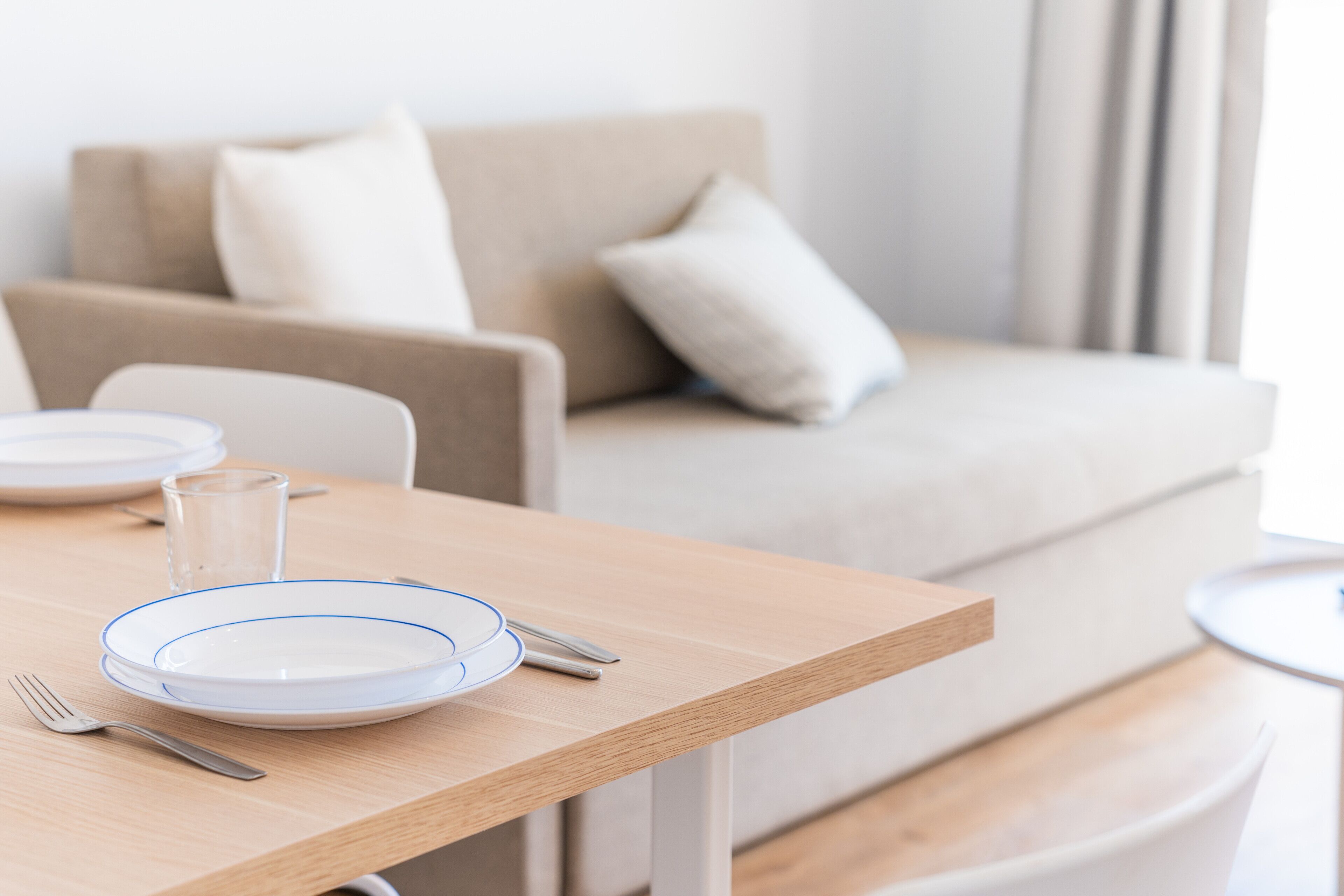 Un comedor con una mesa de madera dispuesta para dos personas, con platos, cubiertos y una copa.