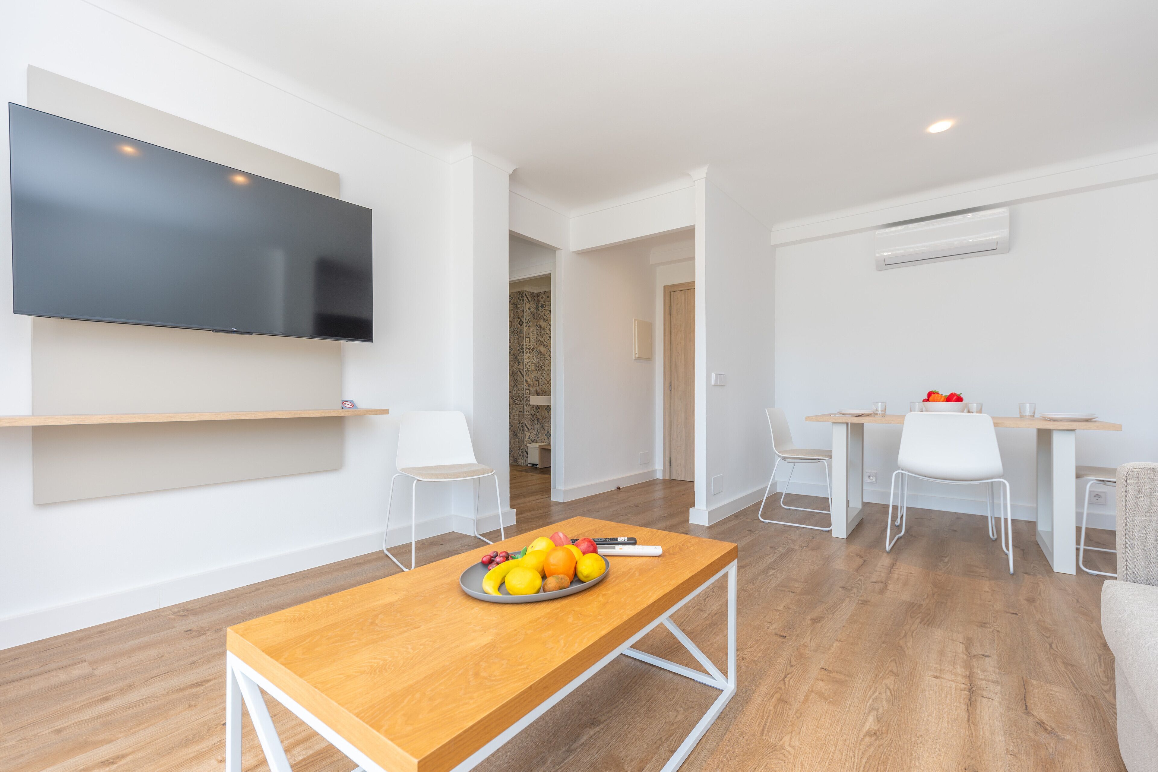 Una sala de estar moderna con un televisor de pantalla plana, una mesa de centro de madera con fruta, sillas blancas y una zona de comedor con mesa y sillas.