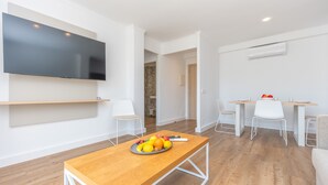 Un soggiorno moderno con una TV a schermo piatto, un tavolino in legno con della frutta, sedie bianche e una zona pranzo con tavolo e sedie.