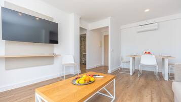 Una sala de estar moderna con un televisor de pantalla plana, una mesa de centro de madera con fruta, sillas blancas y una zona de comedor con mesa y sillas.