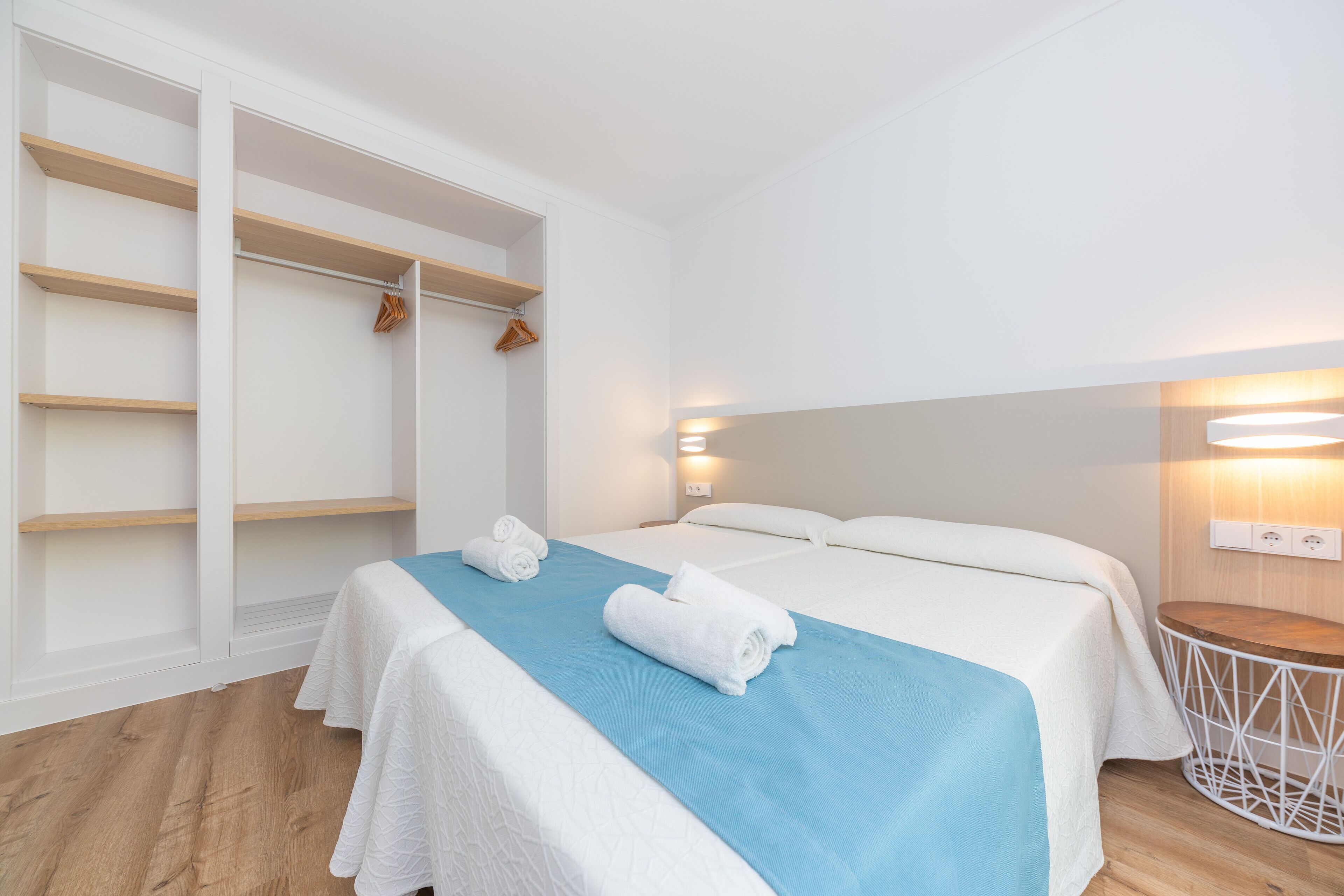 Habitación de hotel con cama, toalla azul, suelo de madera y armario blanco con espacio para colgar.