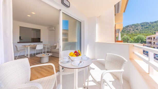 A balcony with white wicker furniture, a table with a fruit bowl, and a view of a hillside.
