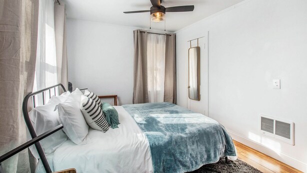 Een slaapkamer met een bed, een plafondventilator, een raam met gordijnen, een spiegel en een wandheater.