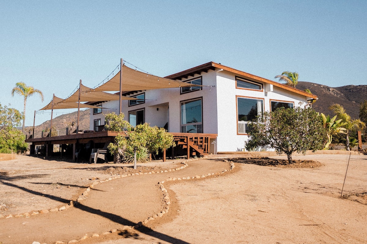A modern house with a large deck, surrounded by desert landscape and palm trees.