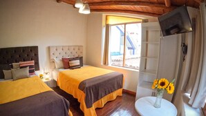 A hotel room with two beds, a small table with a vase of sunflowers, a television on the wall, and a window with a view of a building outside. - Cozy family home in the Sacred Valley (Cusco)
