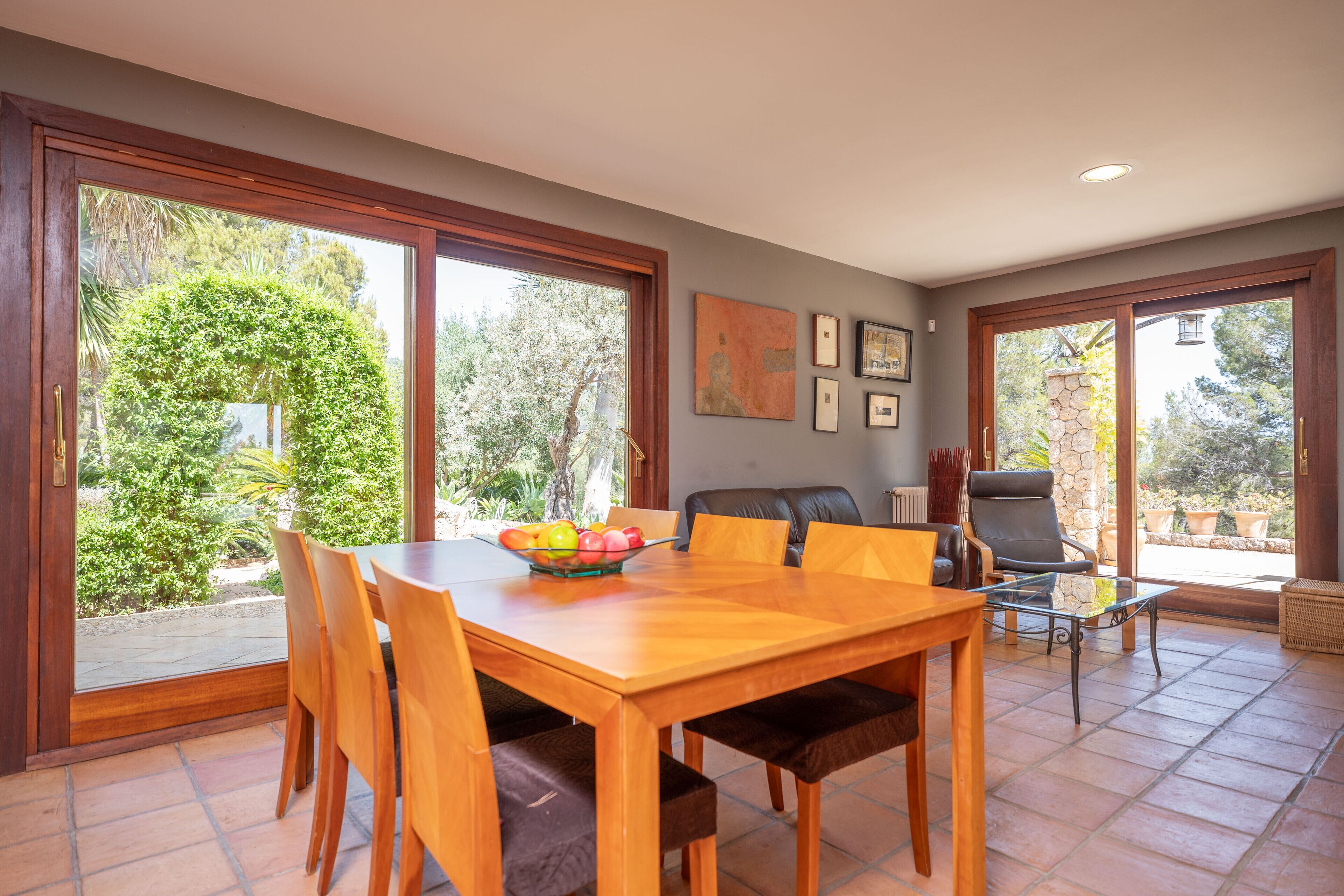 Un coin repas avec une table et des chaises en bois, un panier de fruits et une vue sur le jardin grâce à de grandes portes vitrées.