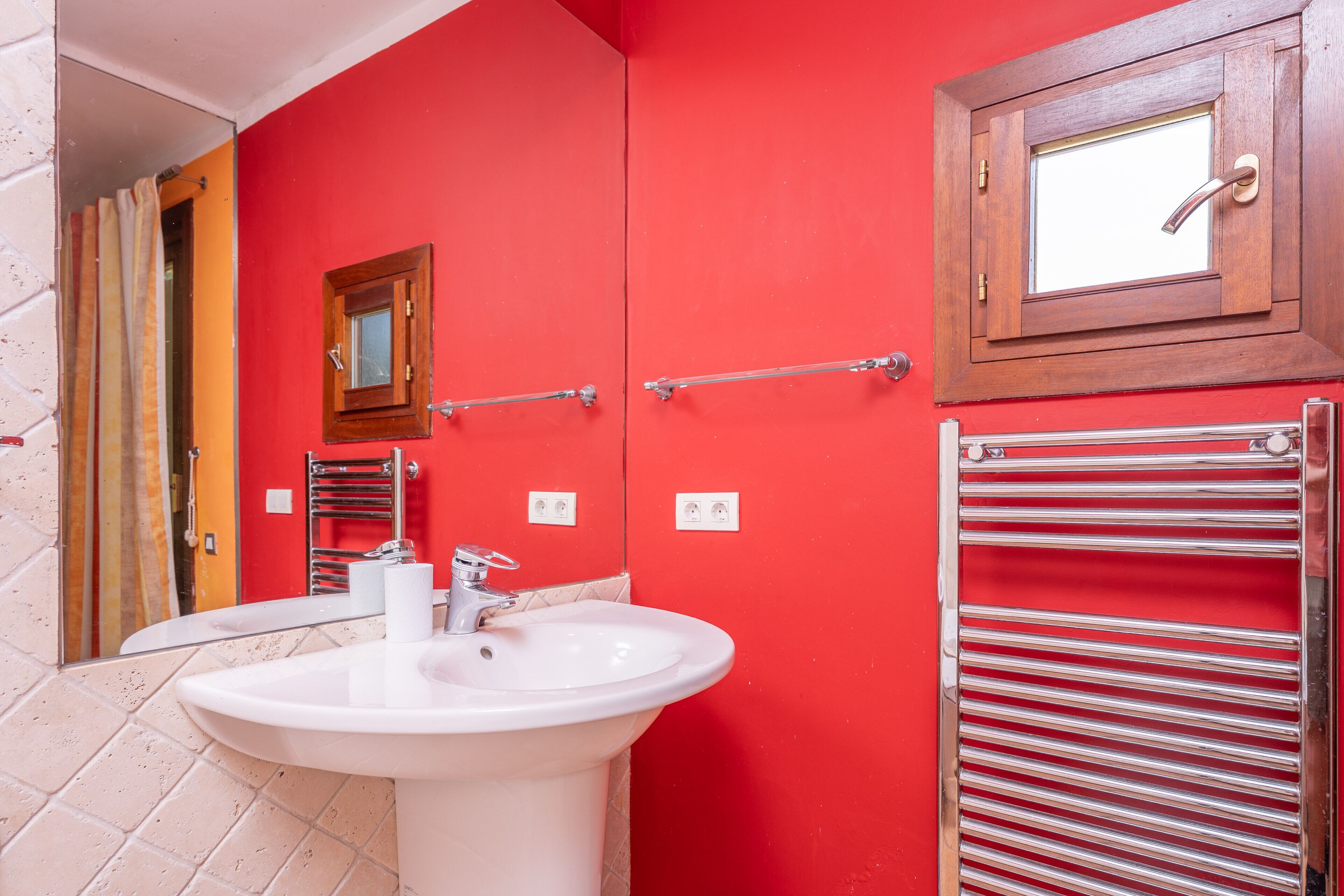 Un baño con una pared roja, un lavamanos blanco, un espejo, un perchero para toallas y una ventana con marco de madera.