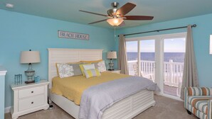 A bedroom with a bed, nightstands, a ceiling fan, a view of the ocean, and a sign that reads 'BEACH HOUSE'.