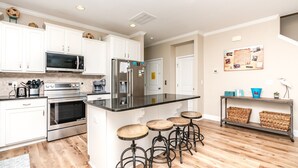 A modern kitchen with white cabinets, stainless steel appliances, a black countertop, and wooden stools. - Crab House - Oceanfront 5 BR Home with Hot Tub & Community Pool (North Topsail Beach)