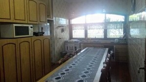 Cozinha com armários de madeira, micro-ondas, uma mesa comprida com toalha de mesa estampada e janela com vista para árvores.