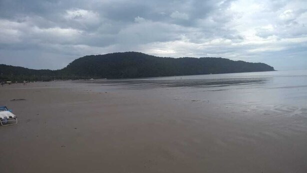 Uma extensa praia de areia com mar calmo e uma colina arborizada ao fundo.
