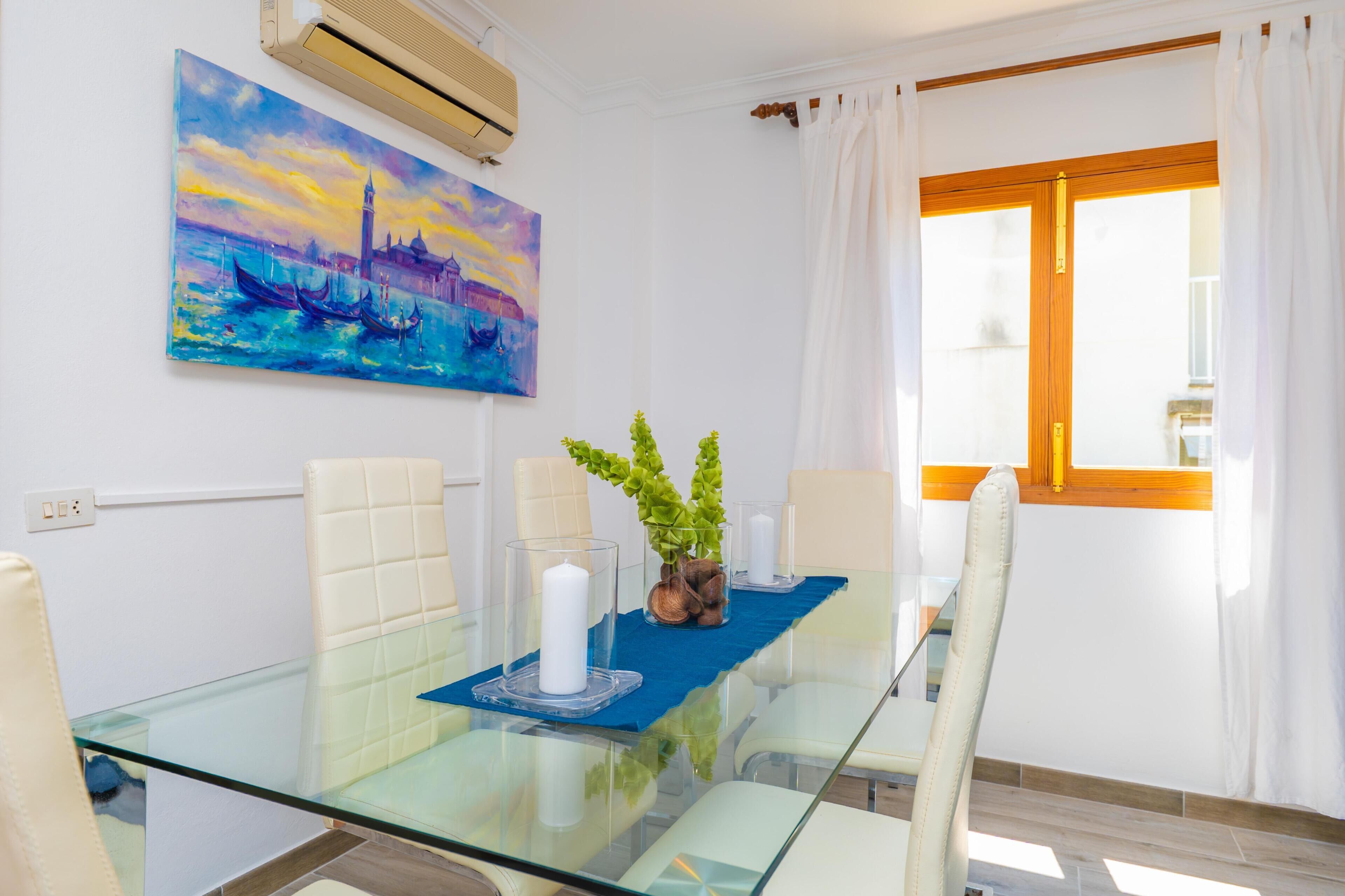 A dining area with a glass table, white chairs, a blue placemat, a white candle, and a painting of a cityscape.