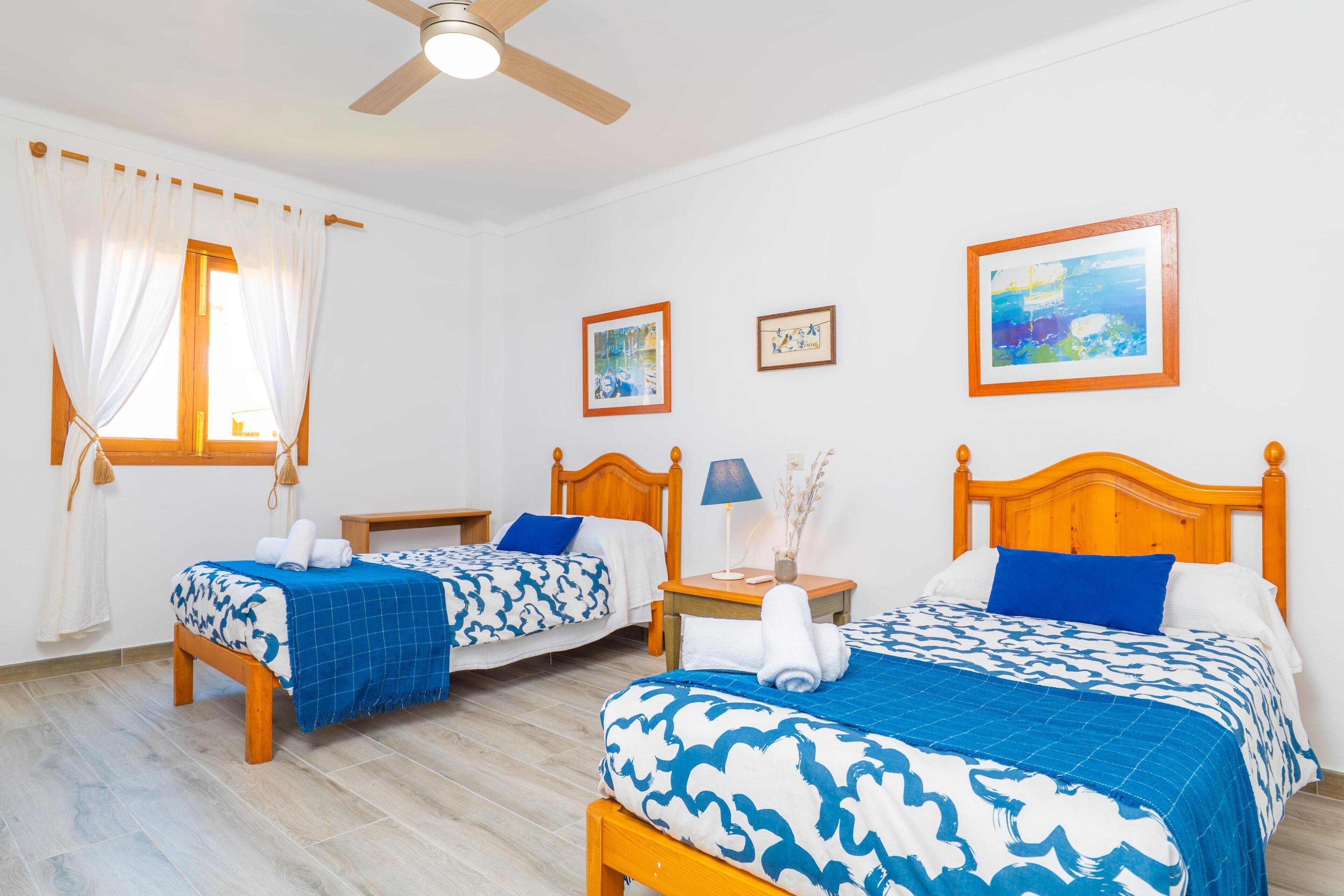 A hotel room with two beds, a ceiling fan, a window with curtains, and artwork on the walls.