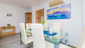 A dining area with a glass table, white chairs, a painting of Venice, a wooden side table, and a wall-mounted air conditioner. - PDM CA'N NOFRE - Fantastic cosy house with terrace and with a the beach just 230 metres aw (Muro)