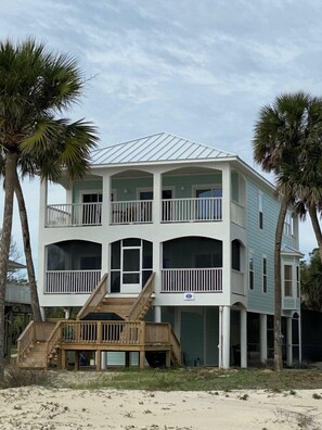 A two-story beachfront house with a balcony, wooden deck, and palm trees.