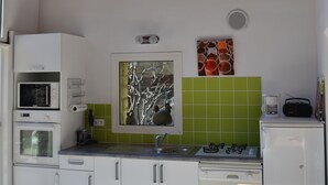 Een keuken met witte kastjes, een magnetron, een fornuis en een spoelbak. Er hangt een klok aan de muur en boven de spoelbak is een raam.