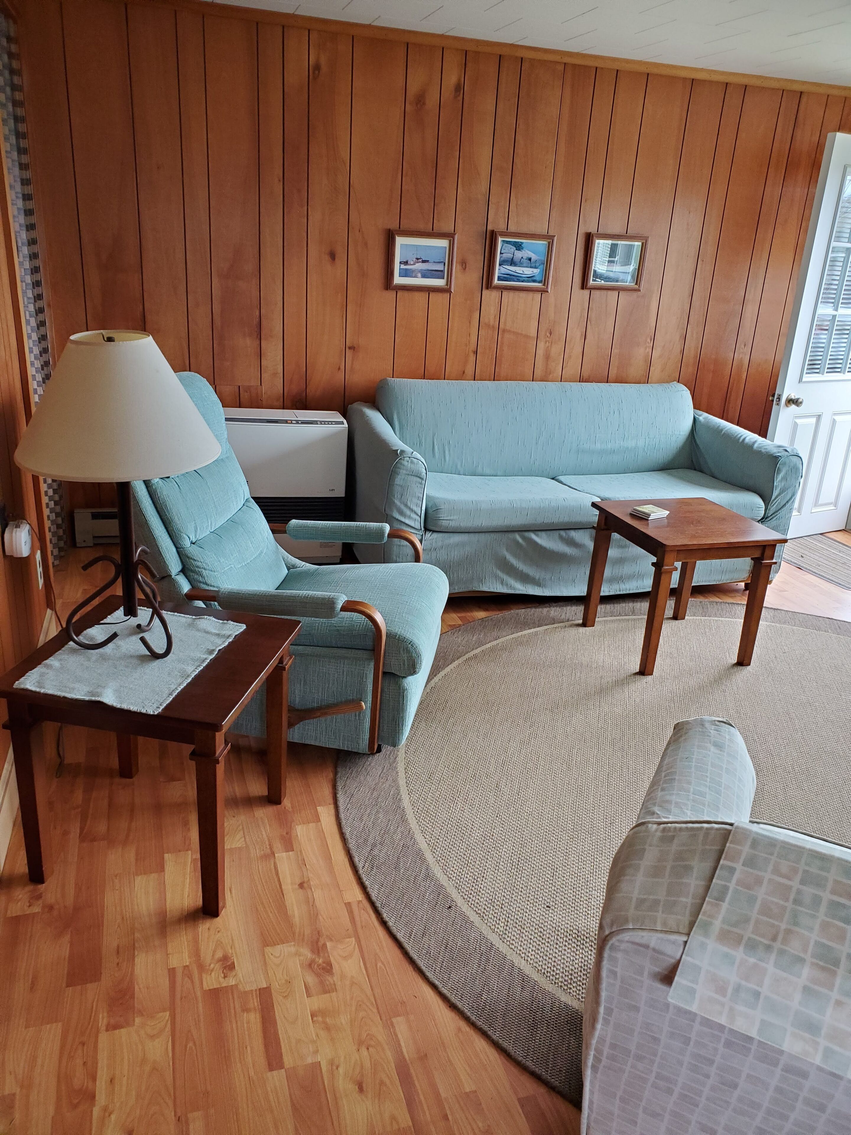 Een gezellige woonkamer met een blauwe bank, een bijpassende fauteuil, een houten salontafel en een vloerlamp. Er hangen ingelijste foto's aan de muur en een raam met zonwering.